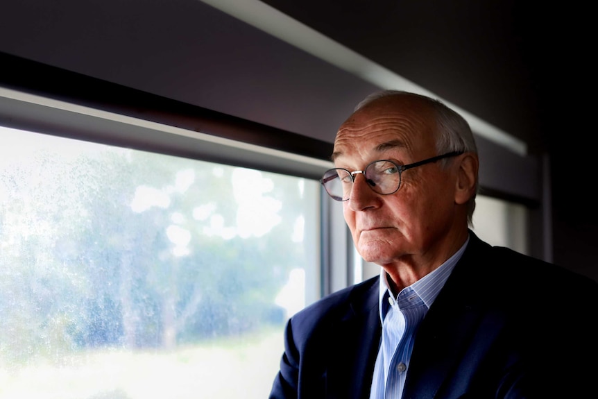 Older man with black rimmed glasses wearing black suit jacket and light-blue striped shirt stands at window