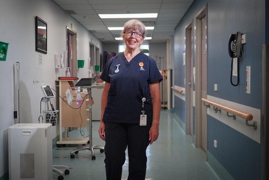A grey haired nurse in scrubs beams in a hospital hallway.