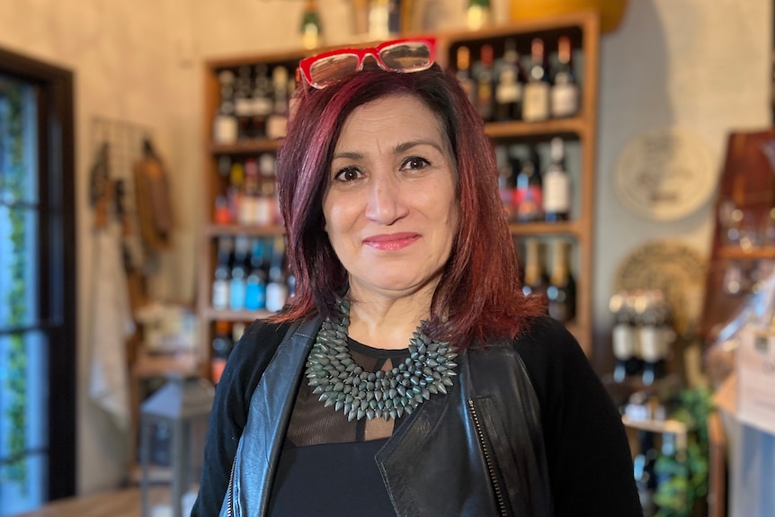 Carmel smiles as she stands inside her warmly lit shop, in front of a shelf stacked with wine bottles.