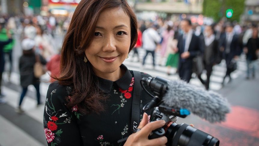 Woman holding a camera standing in the street with crowd of pedestrians walking across a crossing.