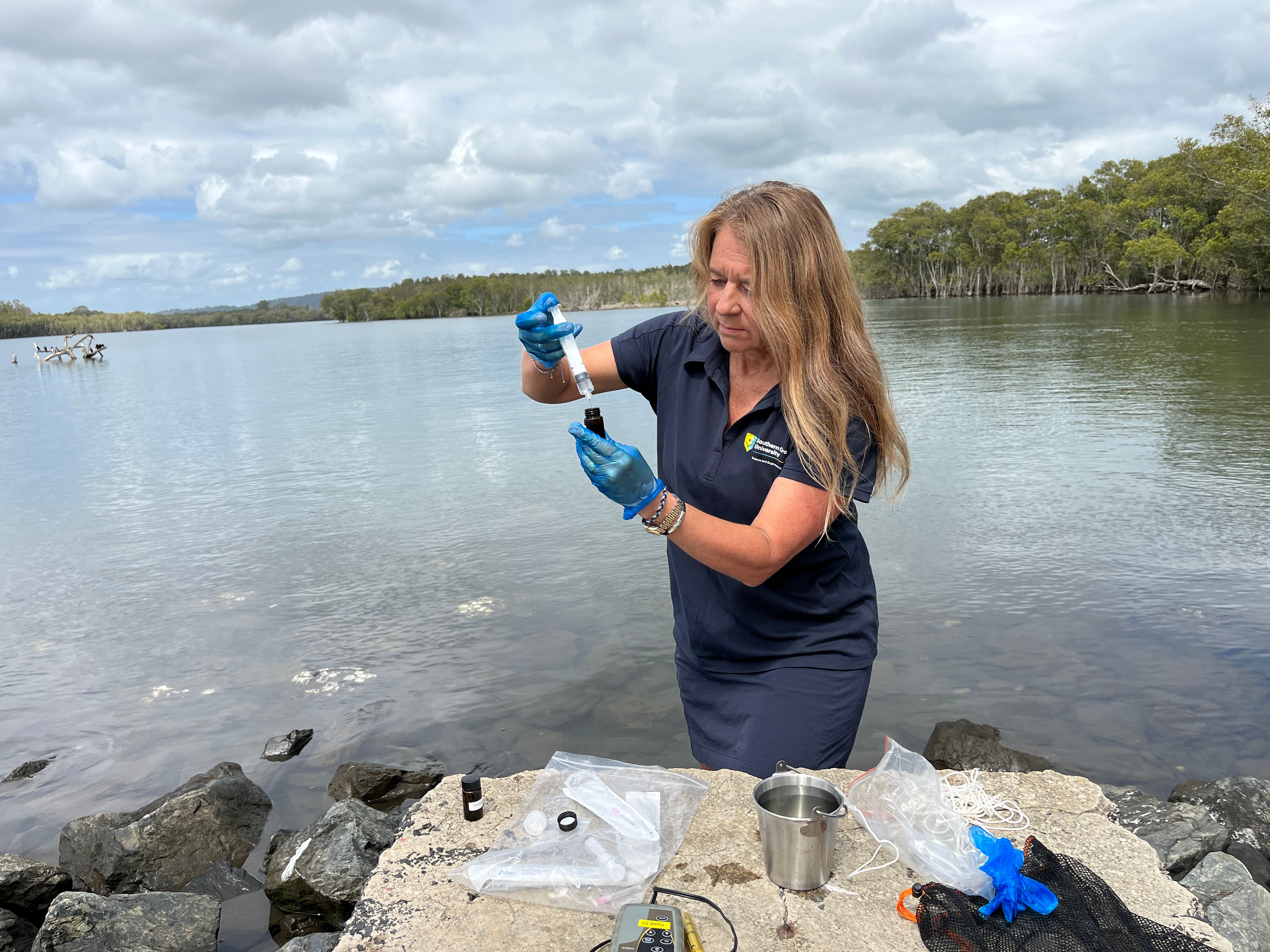 A woman stands on the edge of a river, taking a water sample.