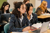 Students wearing pinstriped school uniform jackets looking at laptops and books in a classroom.