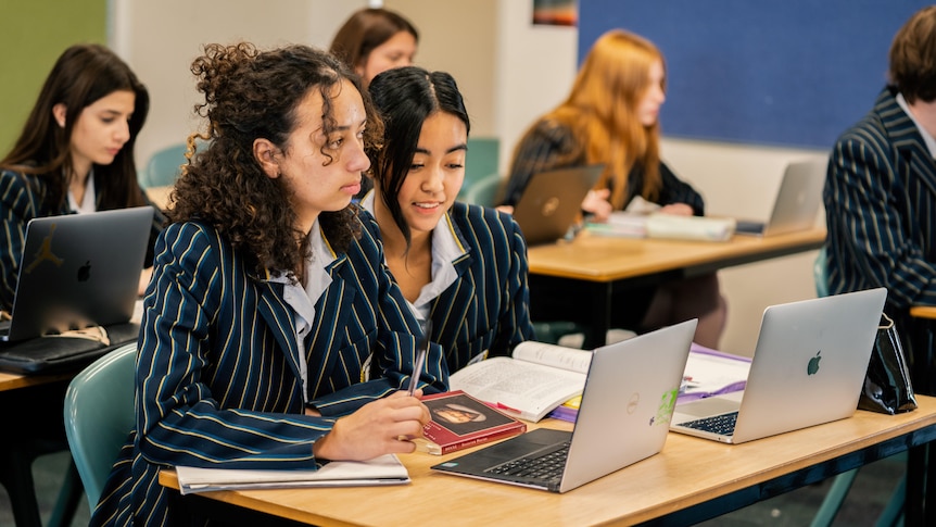 Students wearing pinstriped school uniform jackets looking at laptops and books in a classroom.