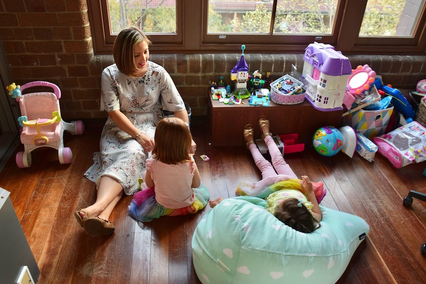 A woman sitting on the floor playing with two young girls surrounded by toys to depict story of life after stillbirth.