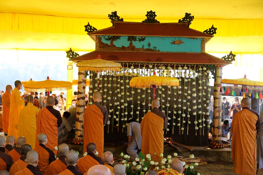 Буддийские монахи в желтых одеждах преклоняют колени вокруг большого украшенного гроба.