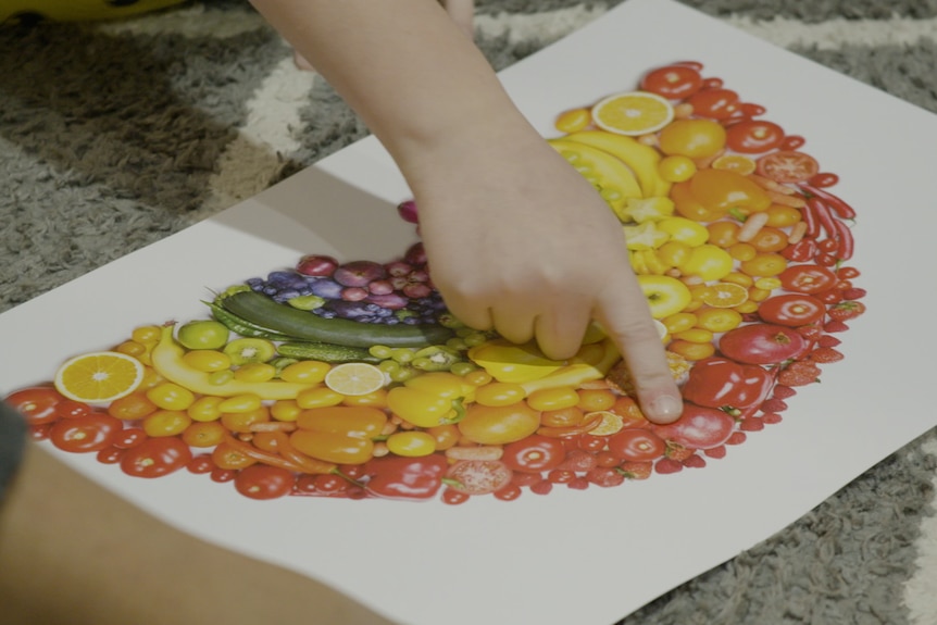 A little boy points at a red apple on a picture of rainbow fruits