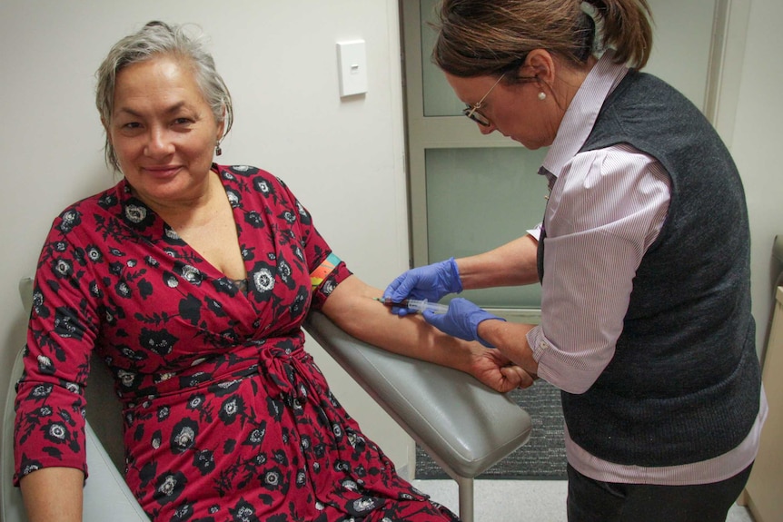 A woman in a colourful dress smiles at camera as she sits in a hospital chair and has an injection into her arm from a nurse.