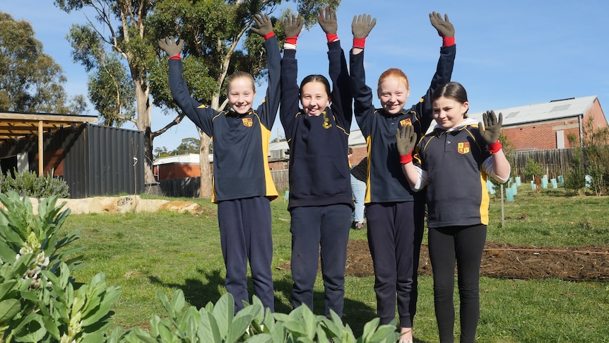 Four primary school girls stand in a garden and smile at the camera as they throw their hands in the air 