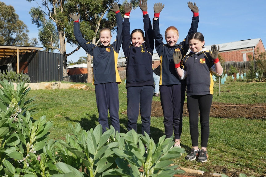 Four primary school girls stand in a garden and smile at the camera as they throw their hands in the air 