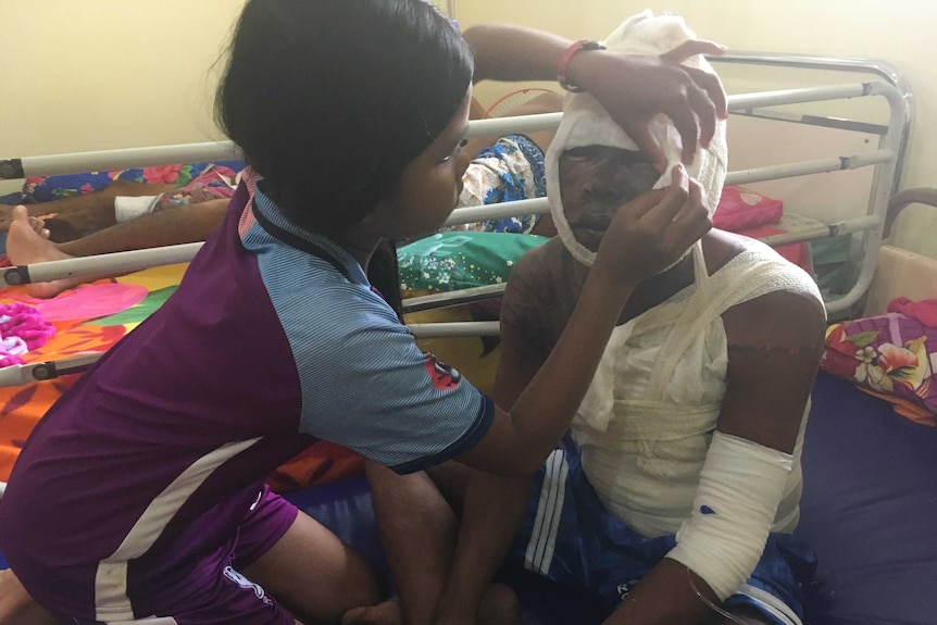 A girl in a purple shirt wipes her father's eyes with a cotton ball on a hospital bed. His head and torso are heavily bandaged.