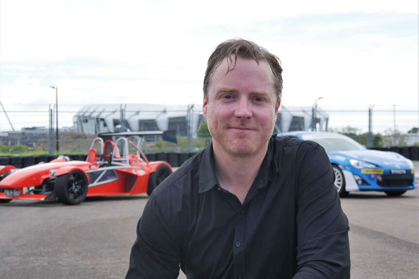 A smiling man on a race track in front of two race cars.
