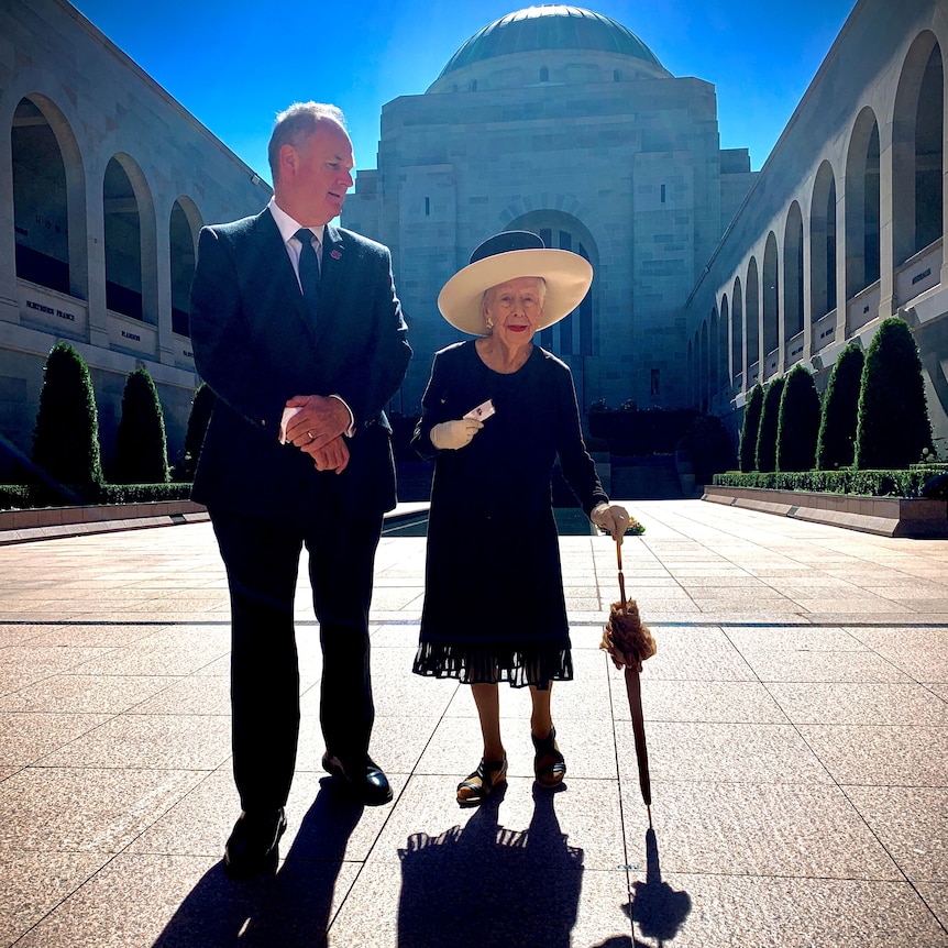 A man guides an older woman in fine clothing through a sandstone courtyard.