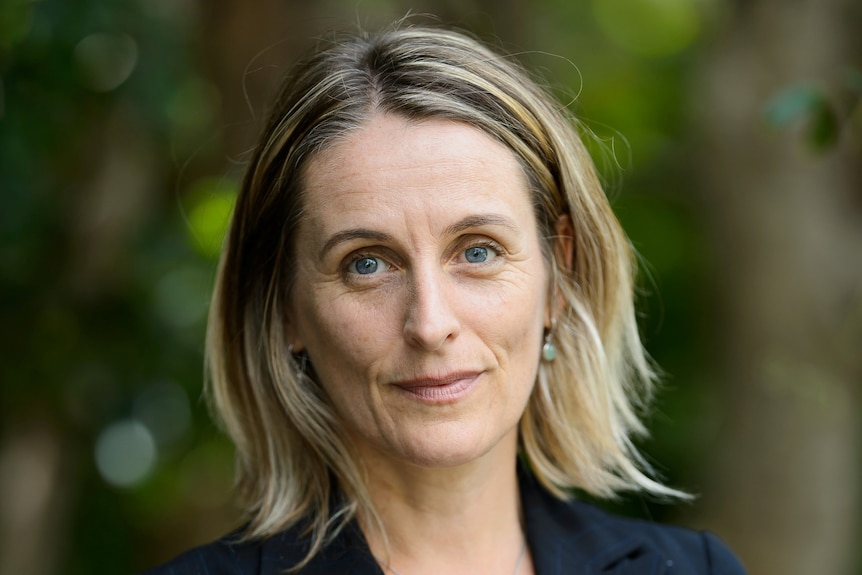 A woman with blonde hair wearing a navy jacket looks at the camera.