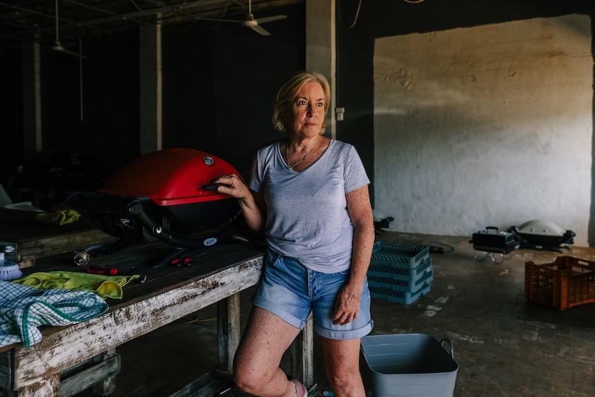 A blonde woman in a tshirt and shorts stands holding a Weber bbq