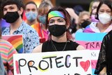 A woman wearing a mask in a crowd of protesters holds a sign that reads "Protect trans kids"