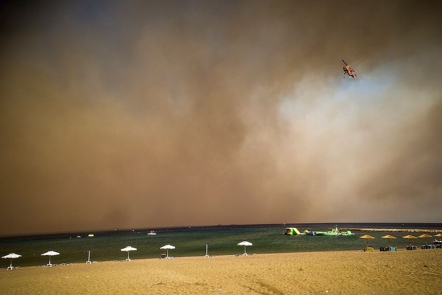 A helicopter hovers over an empty beach on the island of Rhodes with black smoke in the distance.