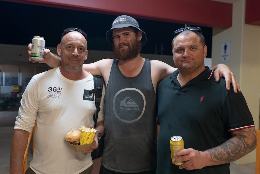 Three men smile at the camera holding beers and food.