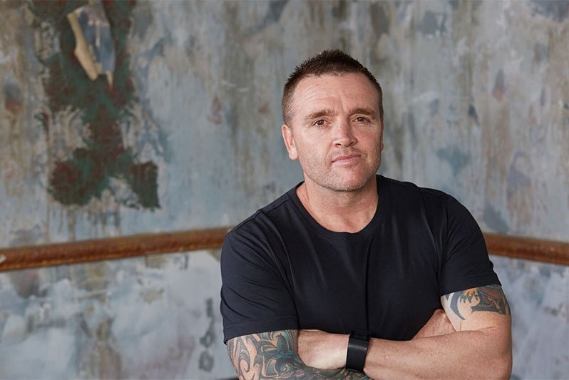 A man with short hair, wearing a black t-shirt, standing with his arms crossed in front of a concrete wall.