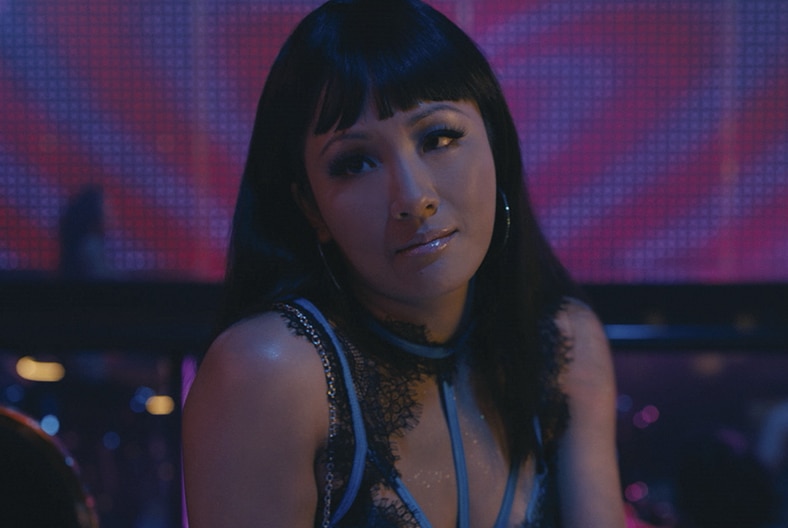 Constance Wu sits wearing hoop earrings and a lacy top in a dimly lit club with a pink and purple LED screen.
