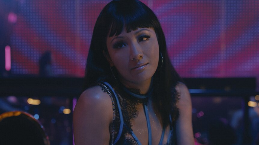Constance Wu sits wearing hoop earrings and a lacy top in a dimly lit club with a pink and purple LED screen.