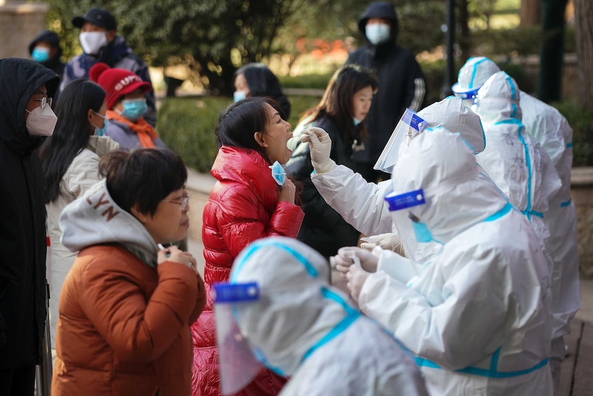 Medical workers in a protective suits take swab from residents, one has their mouth open.