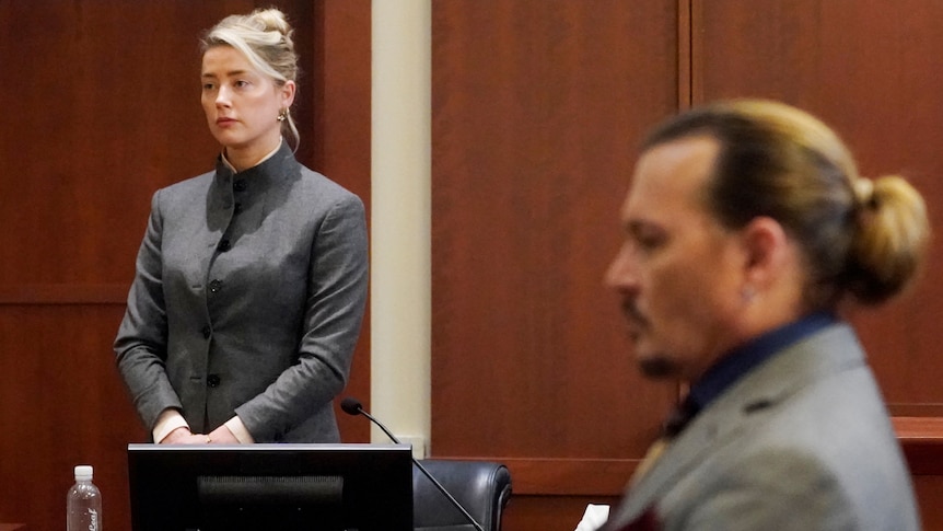 Amber Heard in a grey suit stands in the witness box while Johnny Depp sits in the foreground out of foxu