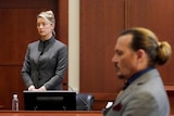 Amber Heard in a grey suit stands in the witness box while Johnny Depp sits in the foreground out of foxu