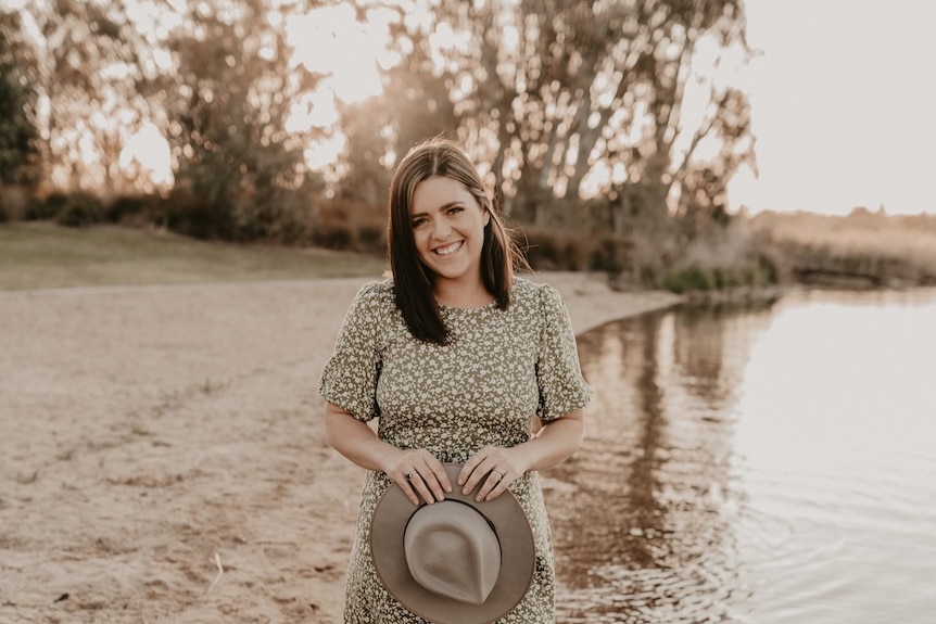 A woman stands on the bank of a river holding a hat and smiling