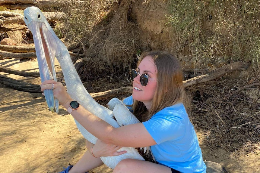 A young woman sits holding a large pelican around the body and by the beak.