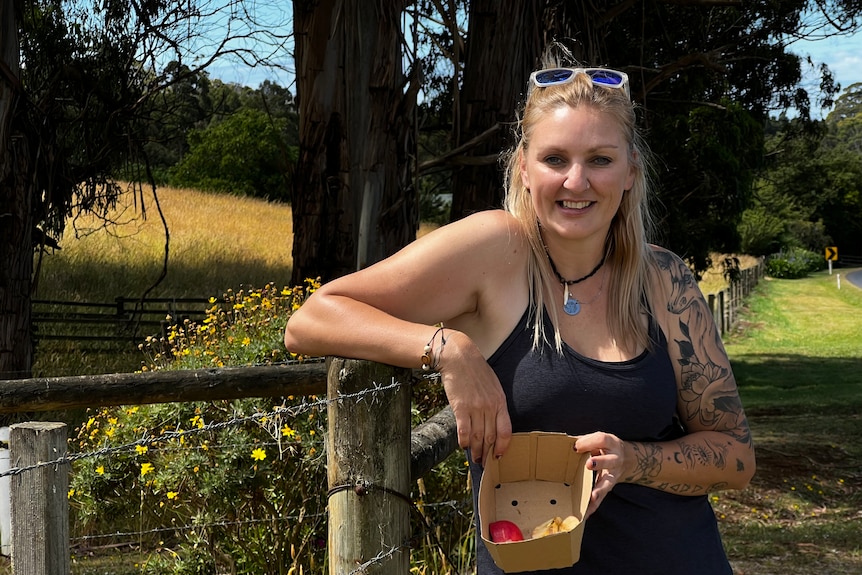 Blonde woman with tattoos and a black top leans up against a paddock fence post.