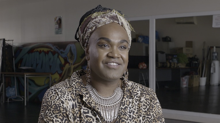 A man wearing head scarf, silver earrings and leopard print top sits smiling in rehearsal room with mirrors during an interview.