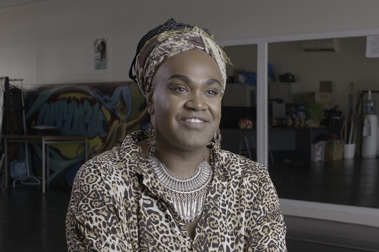 A man wearing head scarf, silver earrings and leopard print top sits smiling in rehearsal room with mirrors during an interview.