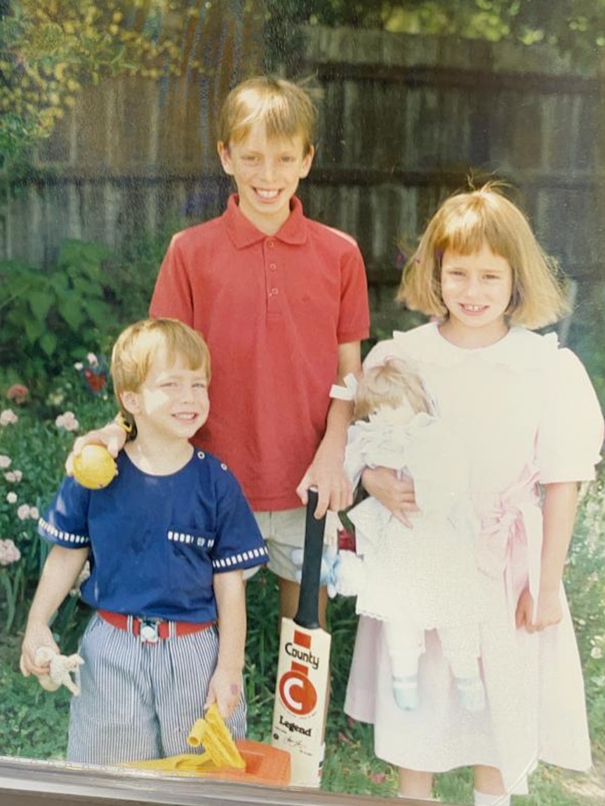 Three kids in the backyard - two boys and one girl. one boy holds a cricket bat