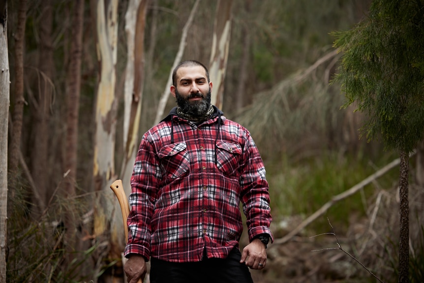 A man in his early 30s, holding an axe and in a red flannel shirt, beard and bald head, standing in Australian bush