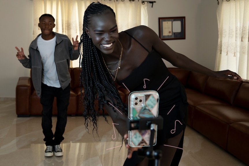 Unice and a friend dance in a living room in front of a mobile phone being held up by a stand.