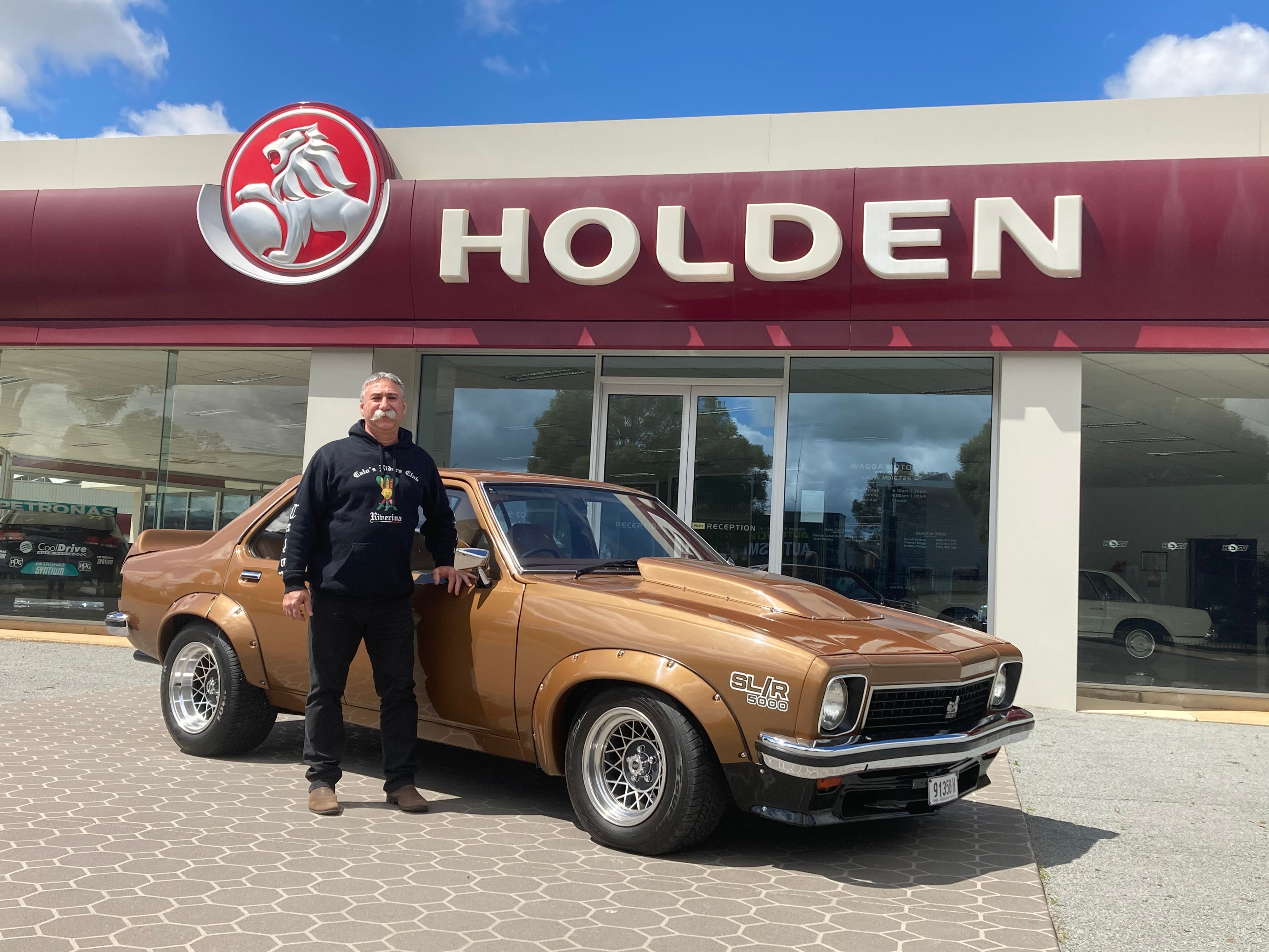 A man in black stands beside a gold coloured sedan.