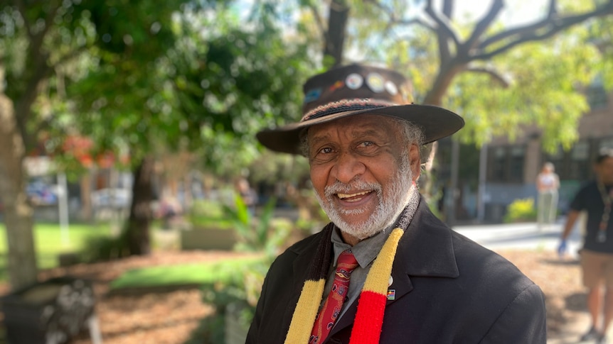 A smiling Indigenous man wearing a hat and scarf stands in a park.