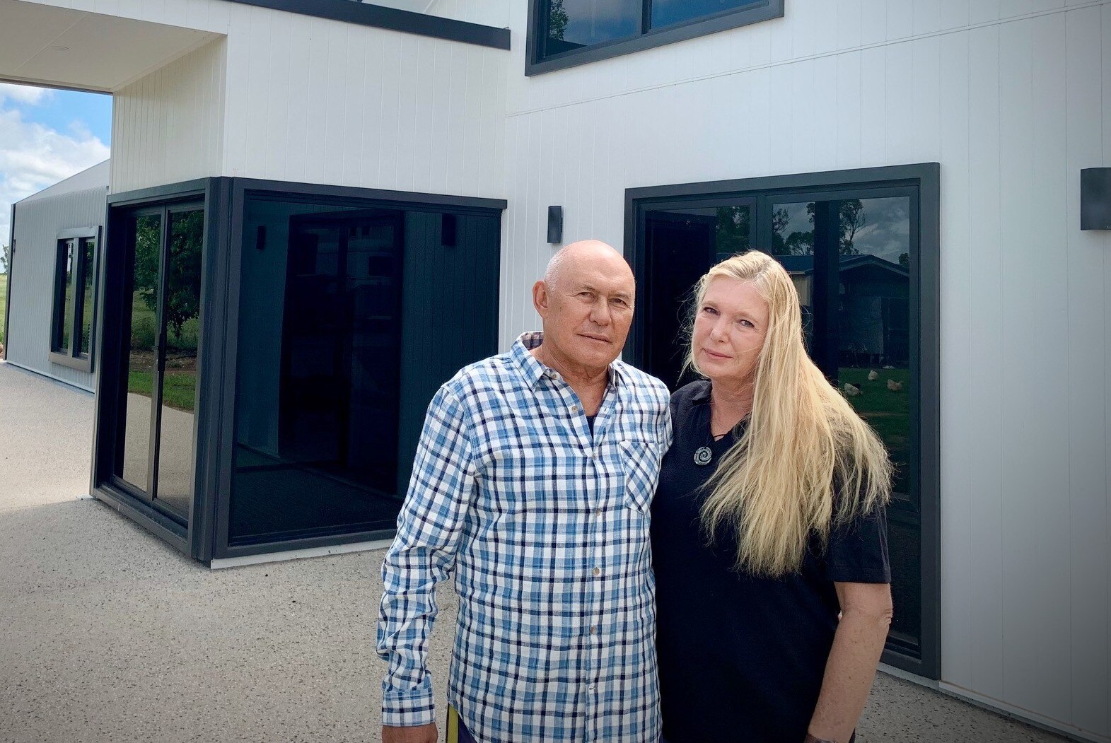 A man in a checked shirt and a woman in a black polo with long blonde hair, in front of a building