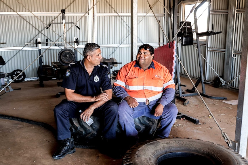 A wide shot of two men sitting on a tyre in a gym, chatting. One is a police officer. The other is wearing high-vis.