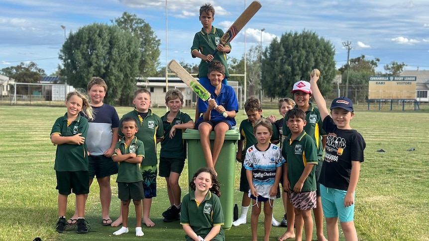 Fourteen kids, some holding cricket bats and some in school uniform, pose for a photo on an oval.