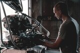 A young man with a shaved head works on an automobile engine in a workshop.
