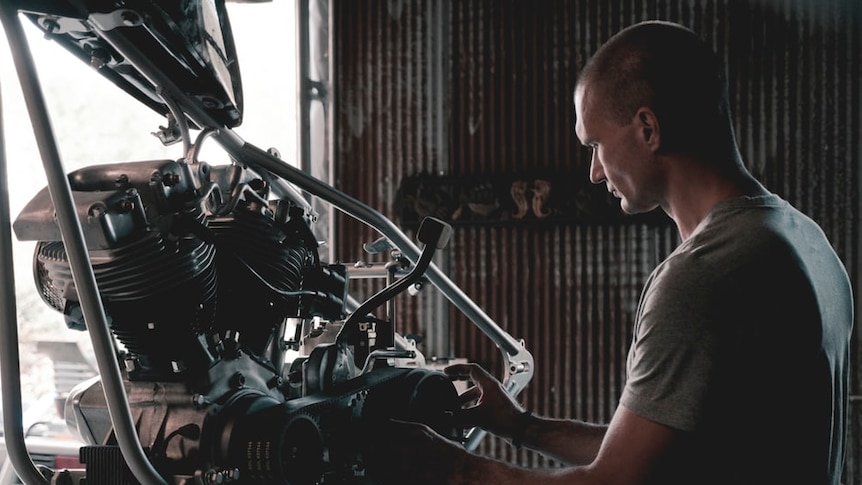 A young man with a shaved head works on an automobile engine in a workshop.
