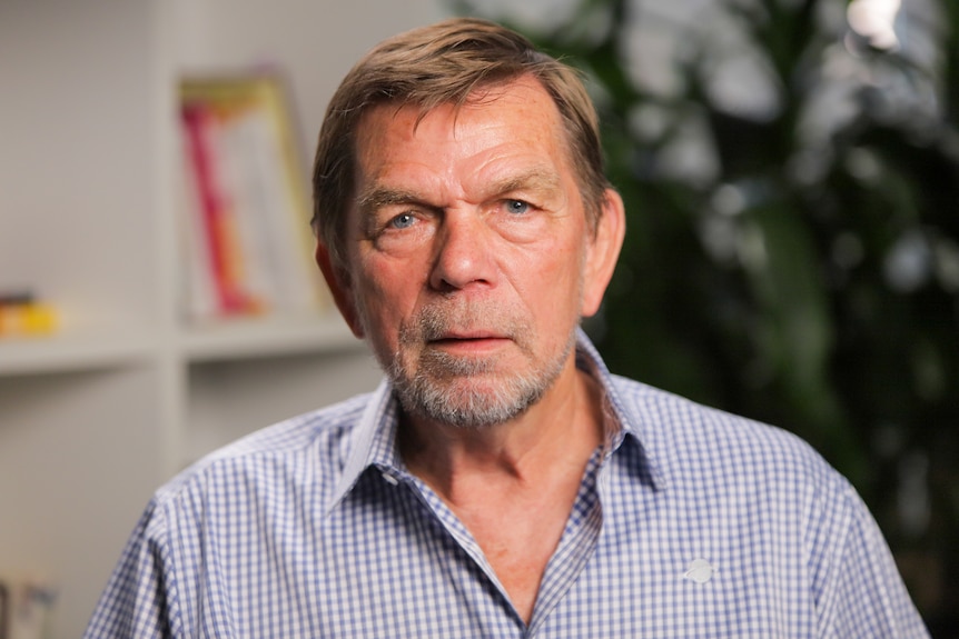 Older man sitting in a travel agency wearing a blue and white checked shirt.