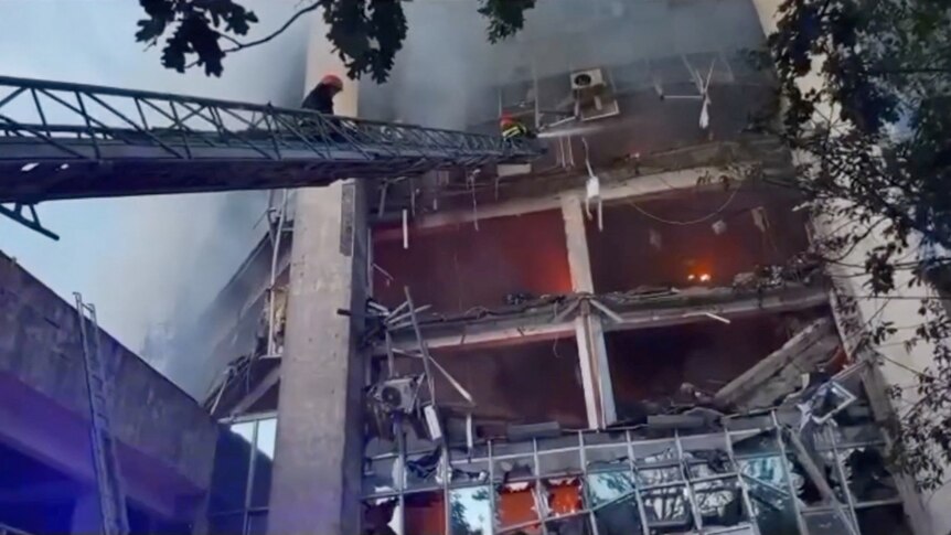 long ladder with two people in hard hats on it, reaching up to a burning and collapsed building