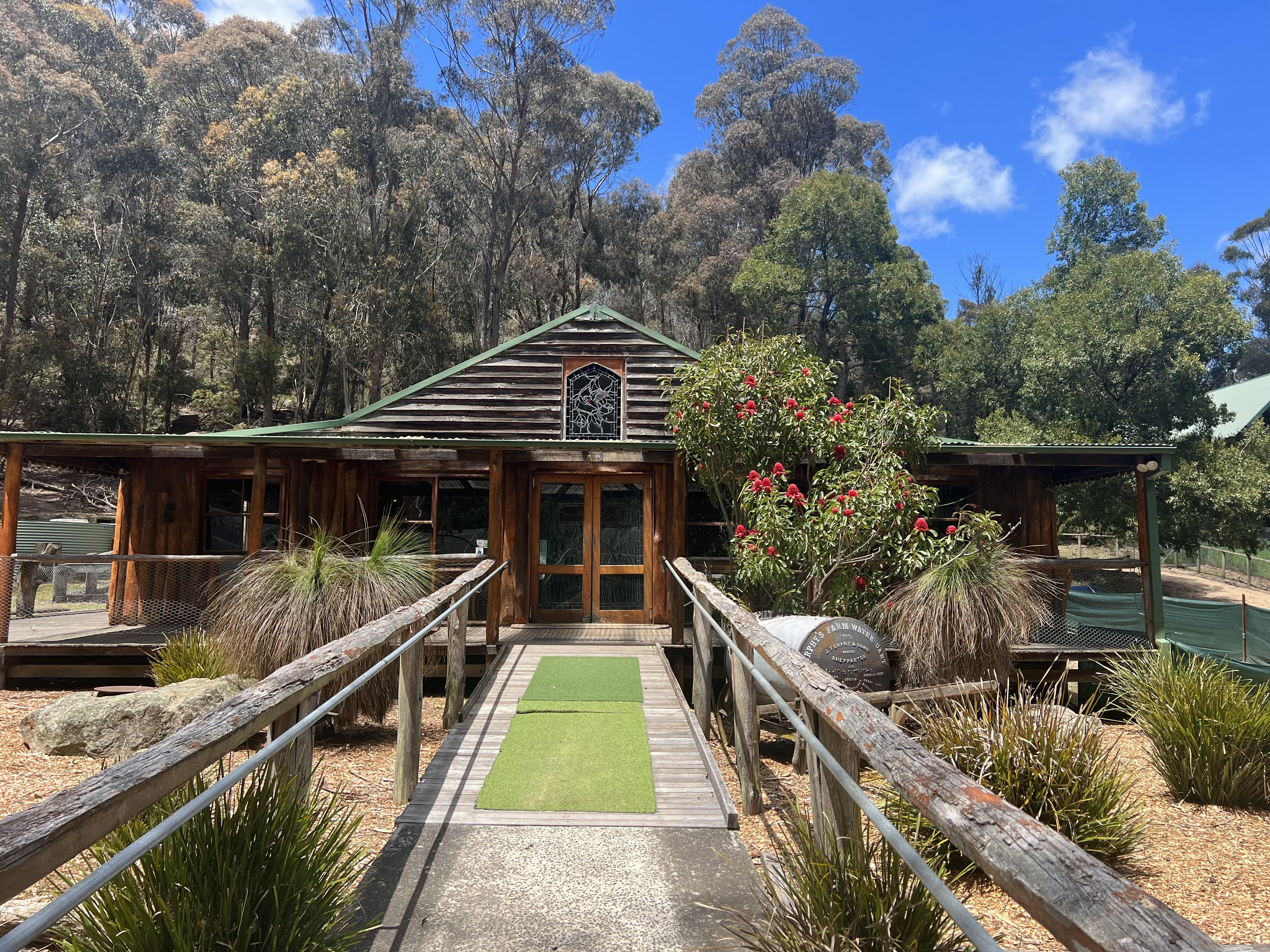 A log house sits on a hillside on a sunny property. Trees and shrubs sit in front of the veranda with trees behind the house.