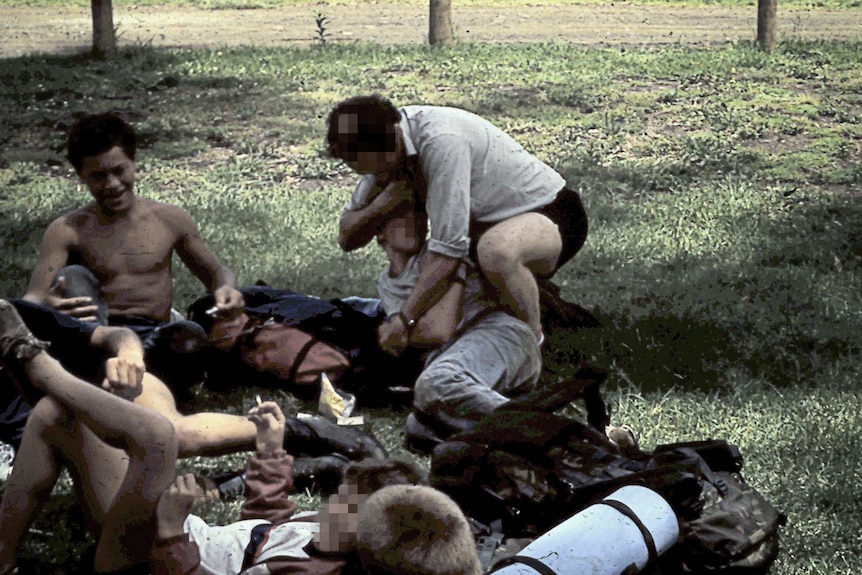 A group of teenage boys sit on the ground, some smoking cigarettes, and a male adult wrestles with one of the boys.