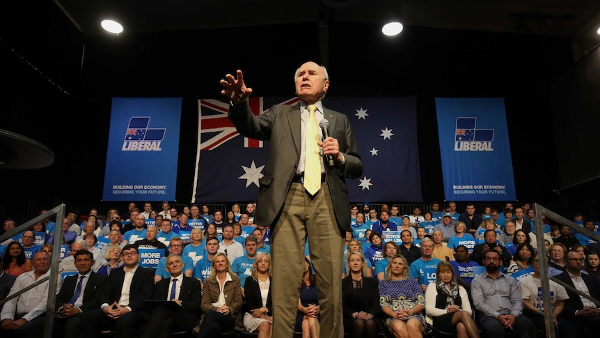 John Howard stands on a stage and reaches out to the crowd. He's surrounded by supporters.