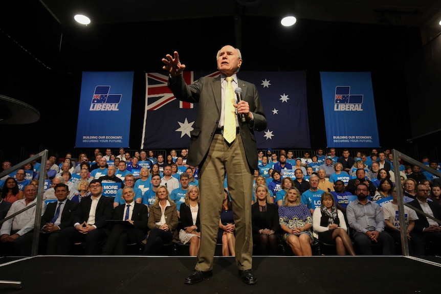 John Howard stands on a stage and reaches out to the crowd. He's surrounded by supporters.