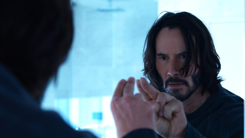 A 57-year-old man with long dark hair and beard looks concerned into a mirror, two fingers probing the surface of the mirror