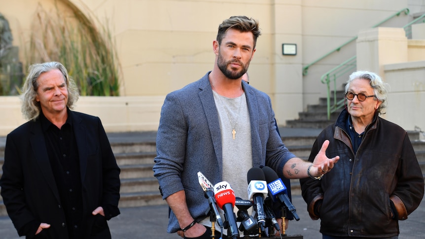 Chris Hemsworth stands at the microphone speaking, two other men stand either side of him